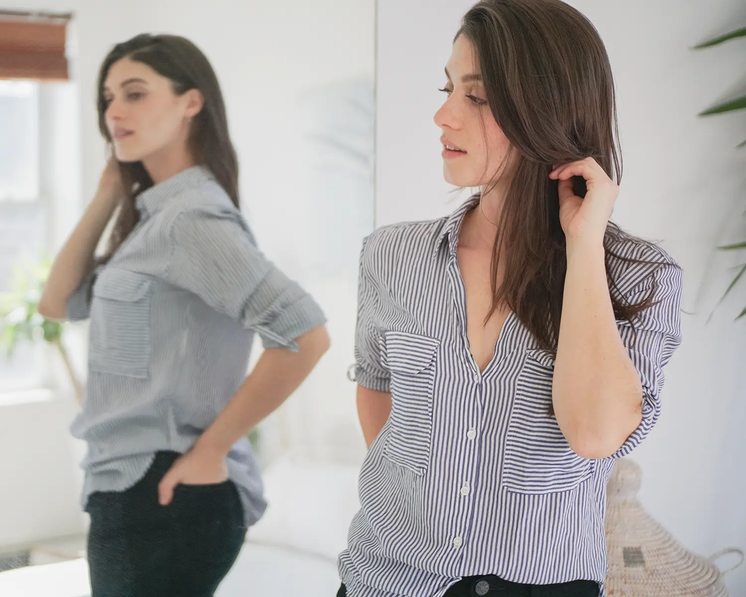 Lifestyle photograph of a woman wearing a striped button-down shirt, shown in motion with her reflection visible in a full-length mirror to illustrate fit
